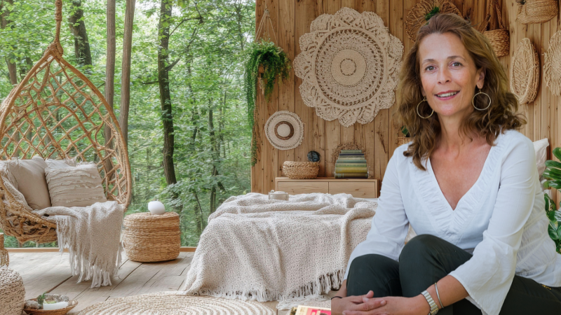 A woman sits in a naturally decorated room with wicker hanging chair and neutral colours, with a floor to ceiling window revealing trees beyond.