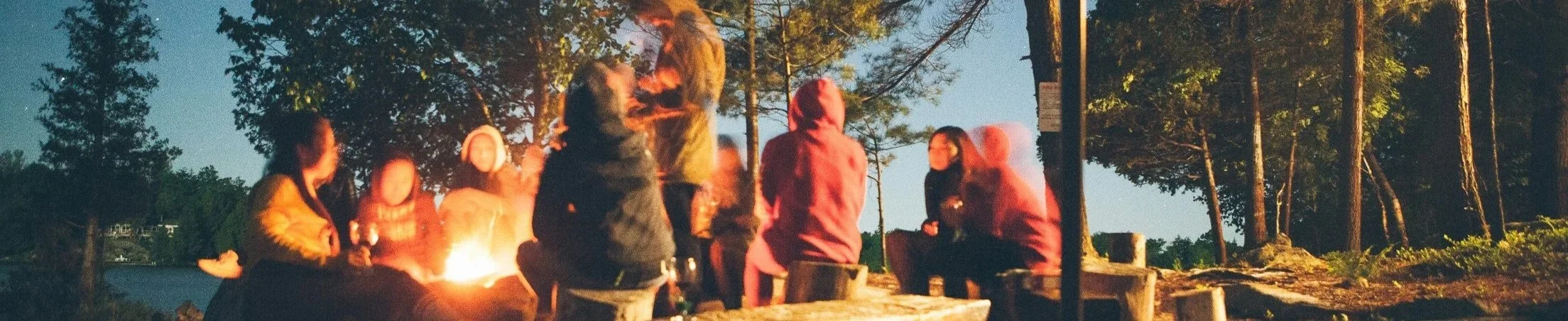 women gather around a campfire. The sky is not yet black and pine trees are visible in the near distance.
