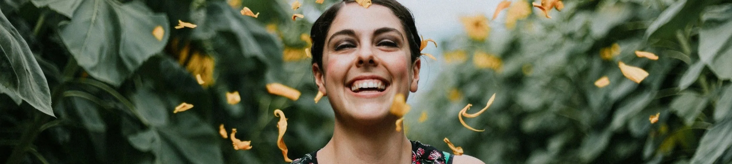 woman stands smiling broadly in a field of sunflowers, petals mid air having been thrown.