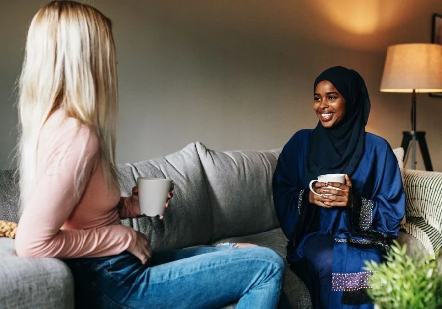 A blonde woman and a dark skinned woman in a hijab chat over coffee on a loungeroom sofa