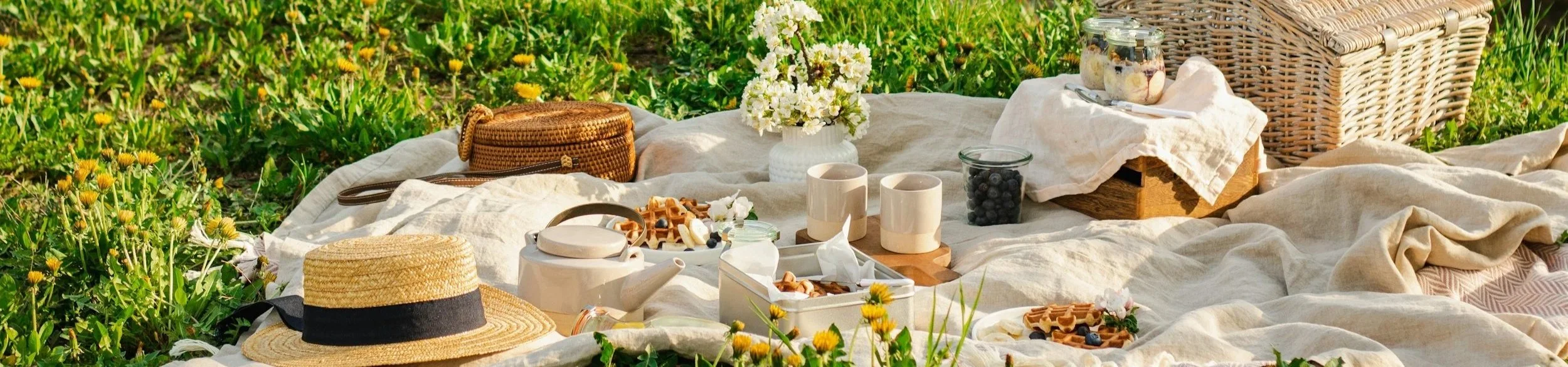 Picnic setting on grass with linen blanket, china mugs and teapot, baskets, food, and straw sun hat in the golden light of late afternoon.