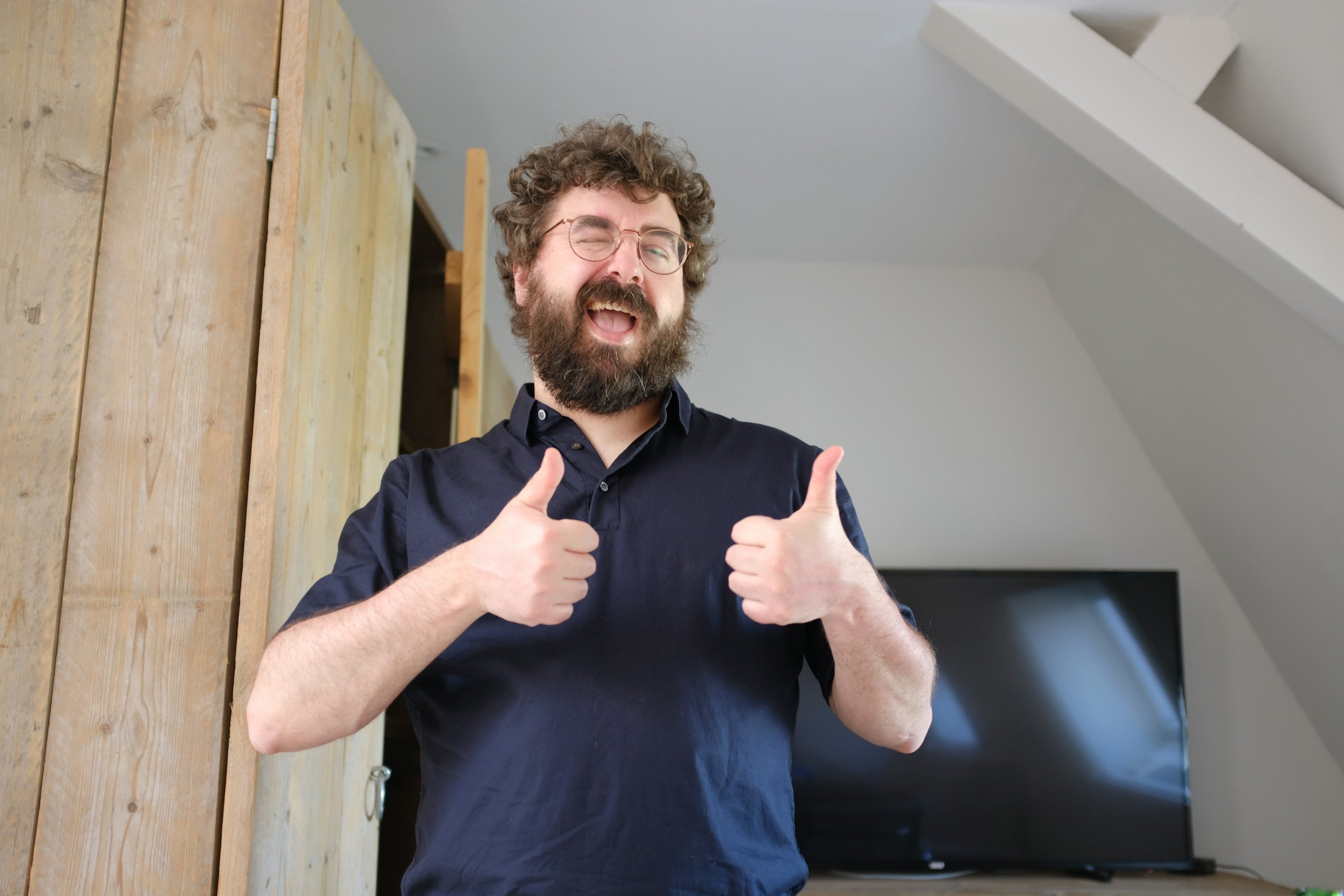 A bearded man with glasses smiles broadly and gives two thumbs up while standing indoors near a wooden door and a flat-screen TV.