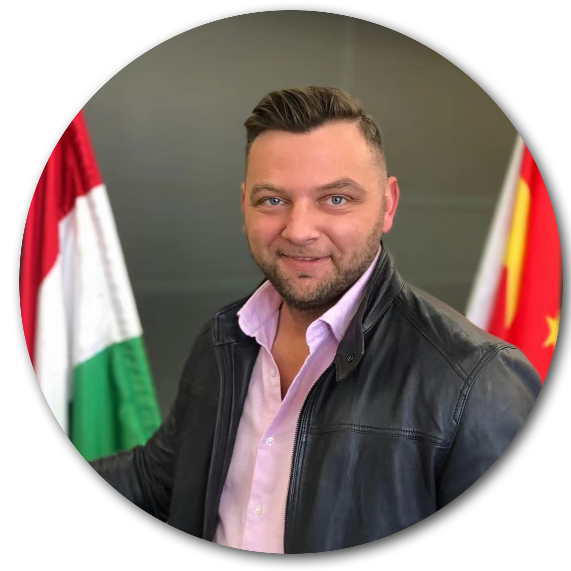 Man in black jacket and pink shirt standing in front of Hungarian and Chinese flags.