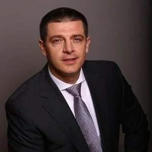 Man in a dark suit and tie posing against a gray background.
