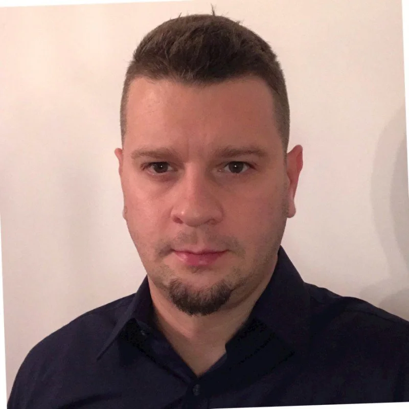 Portrait of a man with short hair, wearing a dark shirt, standing against a plain background.