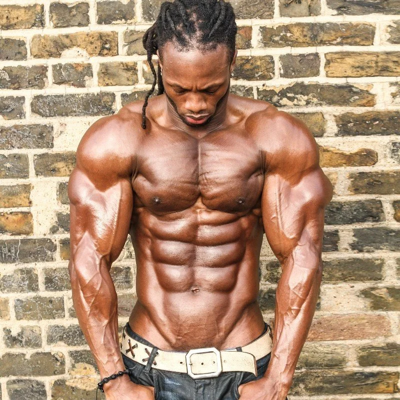 Muscular man with dreadlocks posing shirtless in front of a brick wall, showing defined abs and arms.