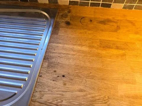 Close-up of scattered black burn marks on an oak kitchen worktop caused by an exploded candle and hot wax fragments.