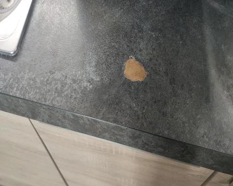 A deep, blistered hot pan burn mark on a dark textured laminate kitchen worktop before repair in Guildford.