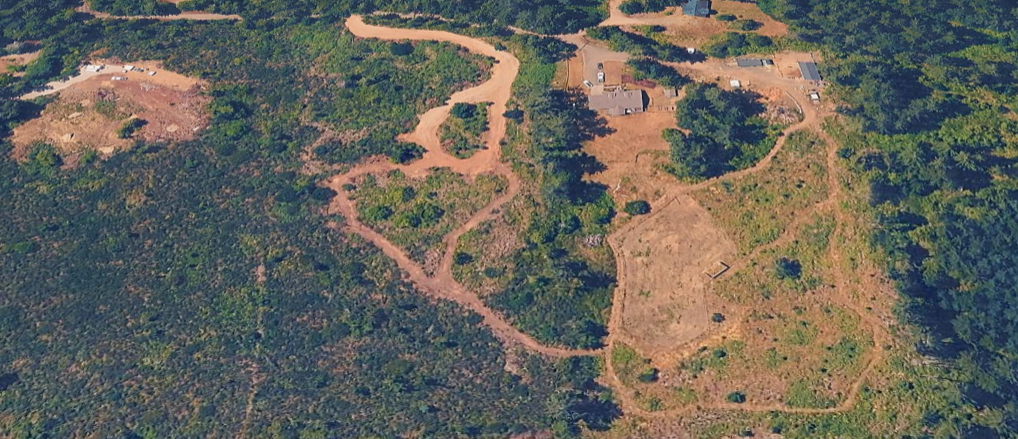 Aerial view of a rural landscape with dirt roads, scattered trees, and a few buildings in the upper right corner.