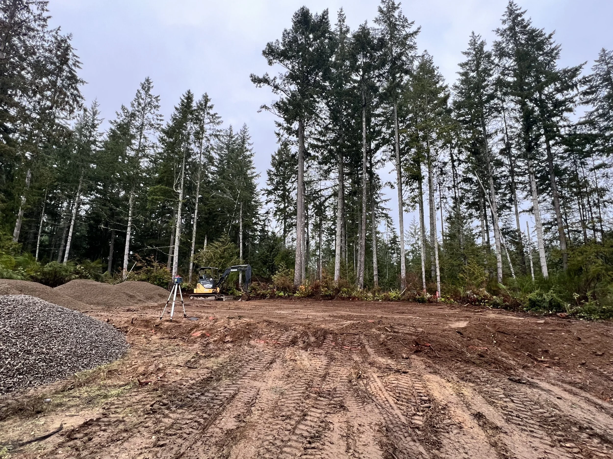 A construction site in a wooded area with tall evergreen trees and some construction equipment, including a small excavator, on a dirt ground with tire tracks and gravel piles.