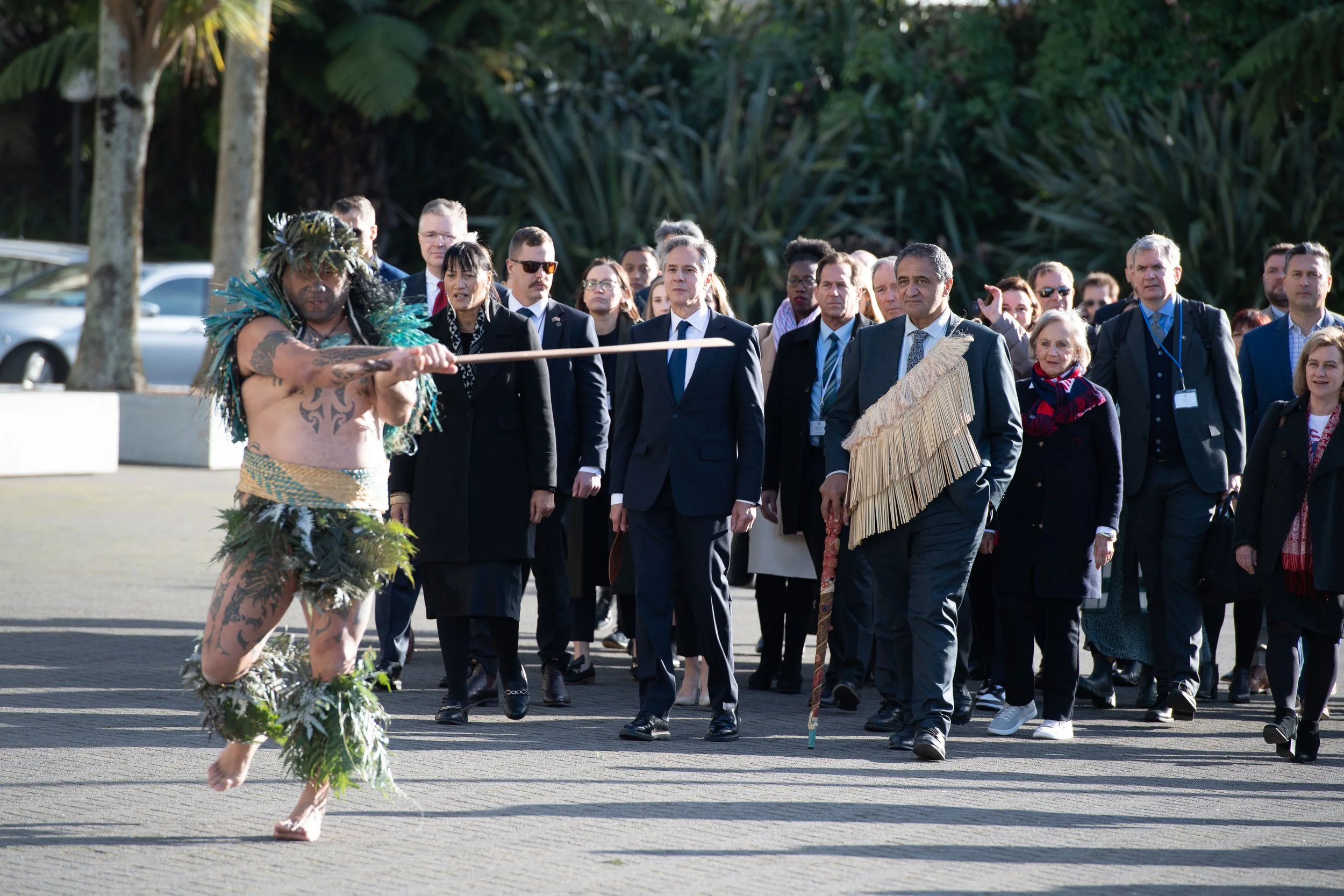 A man in traditional Māori ceremonial attire leads a pōwhiri. He is holding a taiaha (stick) out to the side, while a large group of people in suits walk behind him.