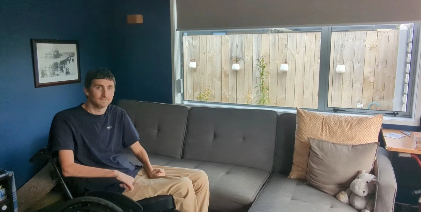 Tim sits in his wheelchair and looks  at the camera. Behind him the blinds in his room are halfway down.
