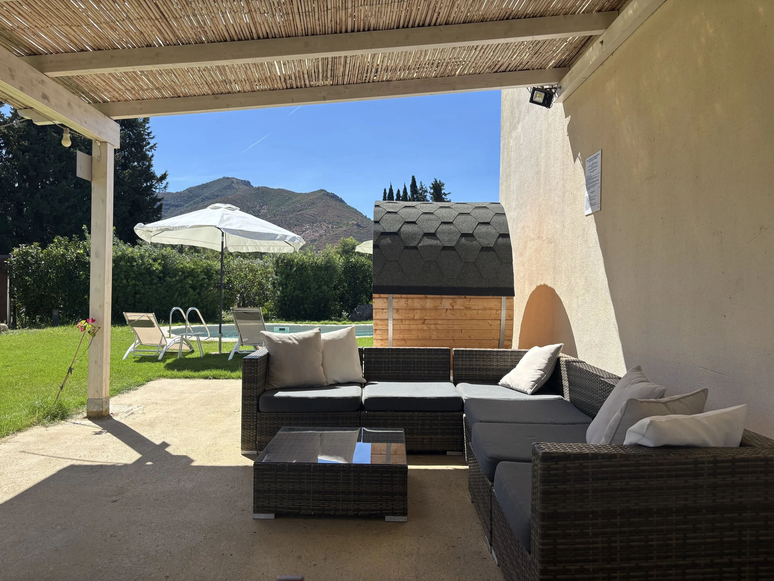 Outdoor patio area with black wicker sectional sofa with white cushions, glass-top coffee table, and shaded sitting area. In the background, there are lounge chairs with umbrellas near a swimming pool, green lawn, trees, and mountains under a blue sk