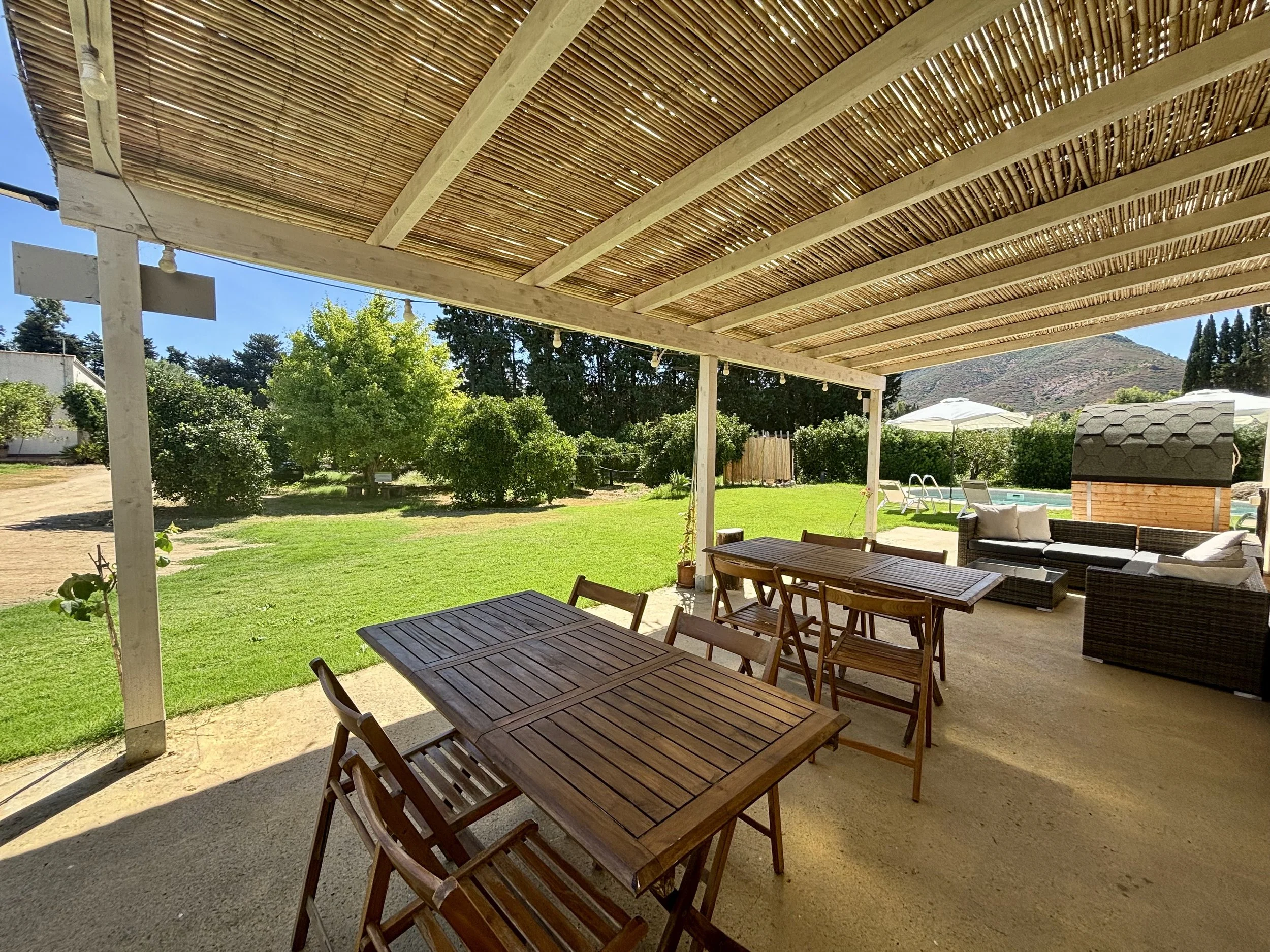 Backyard patio with wooden dining table and chairs, outdoor sofa, shaded pergola, grassy lawn, trees, and clear blue sky.