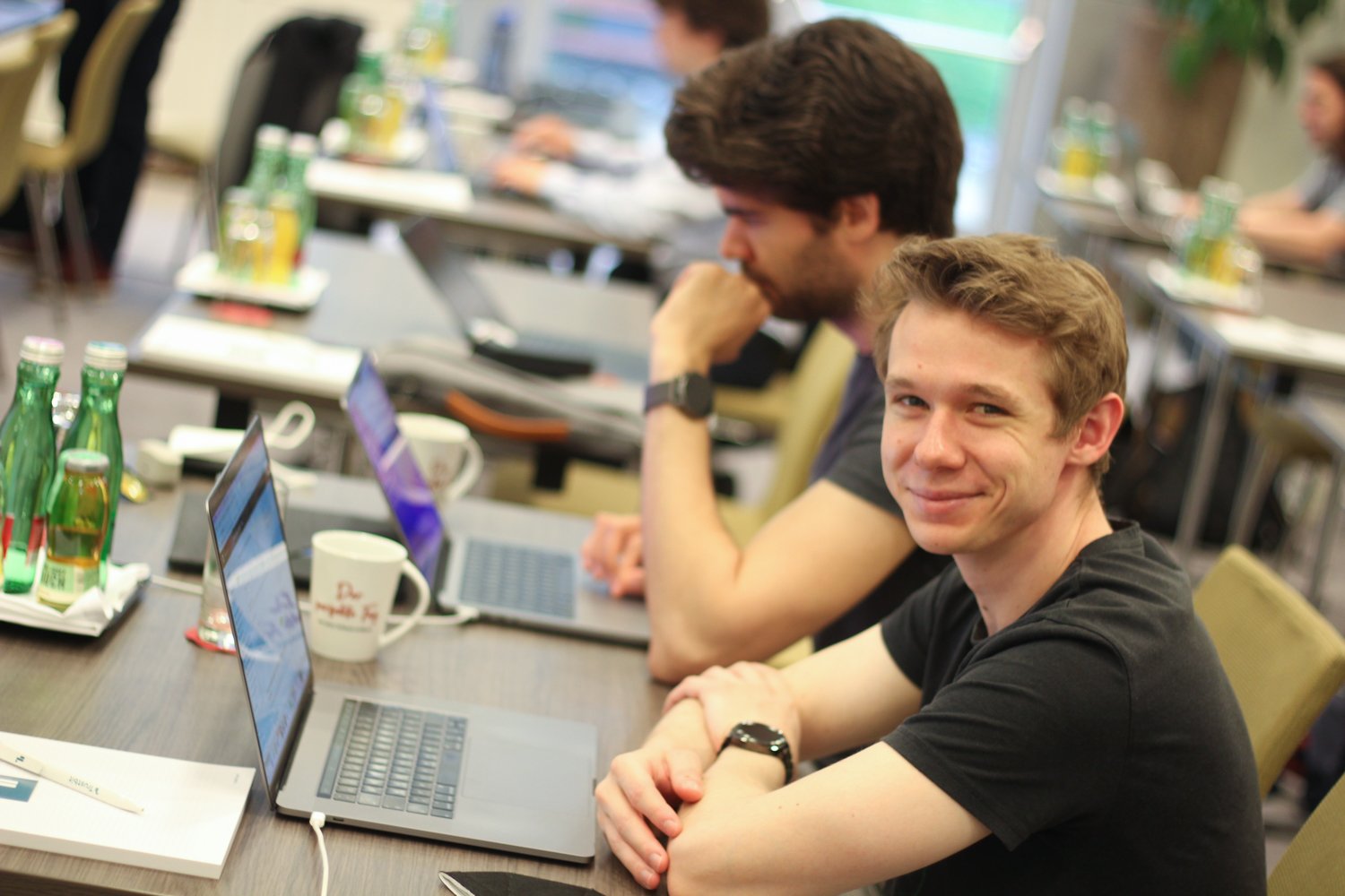 Markus smiling at the camera, sitting at his laptop next to Oscar during a hackathon