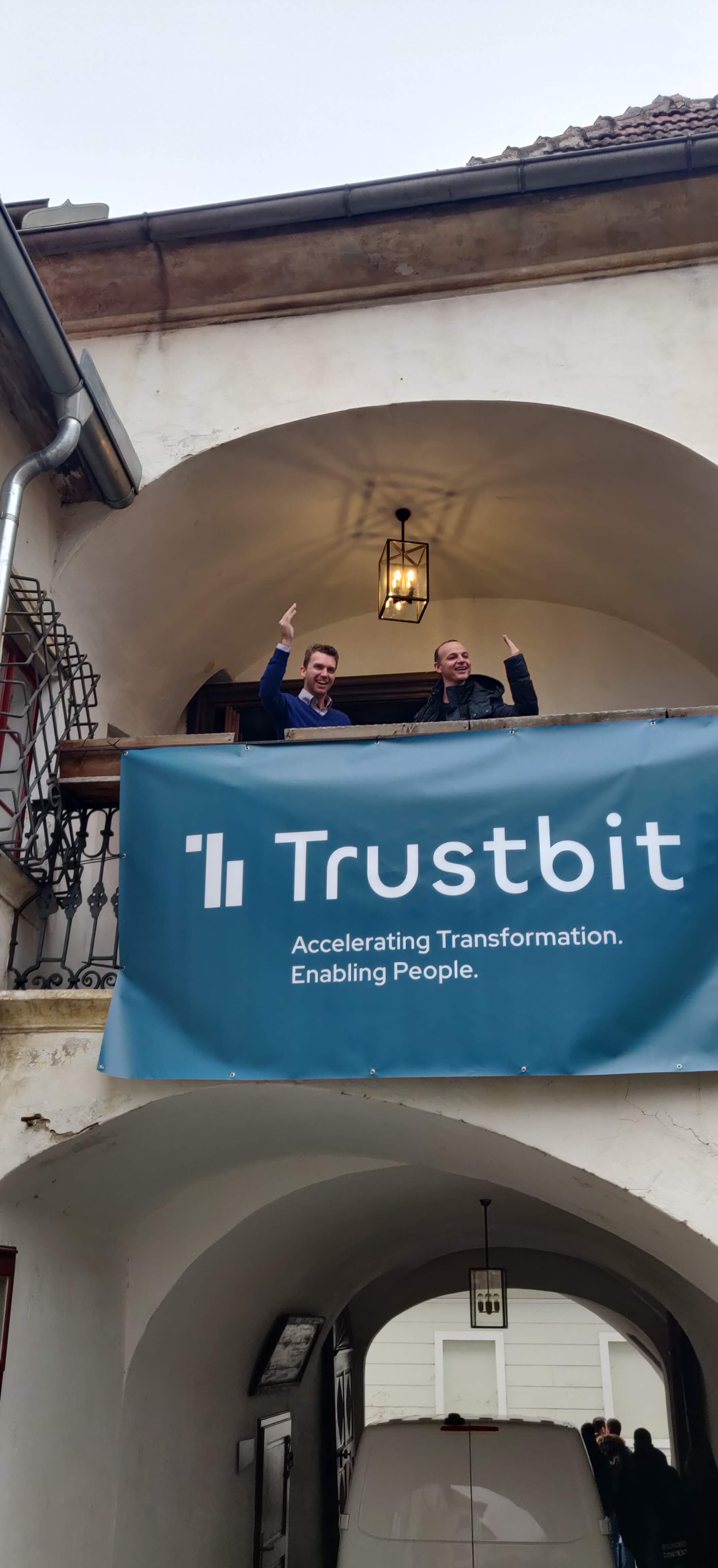 Gottfried and Jörg waving from the first floor balcony of a building behind a Trustbit banner