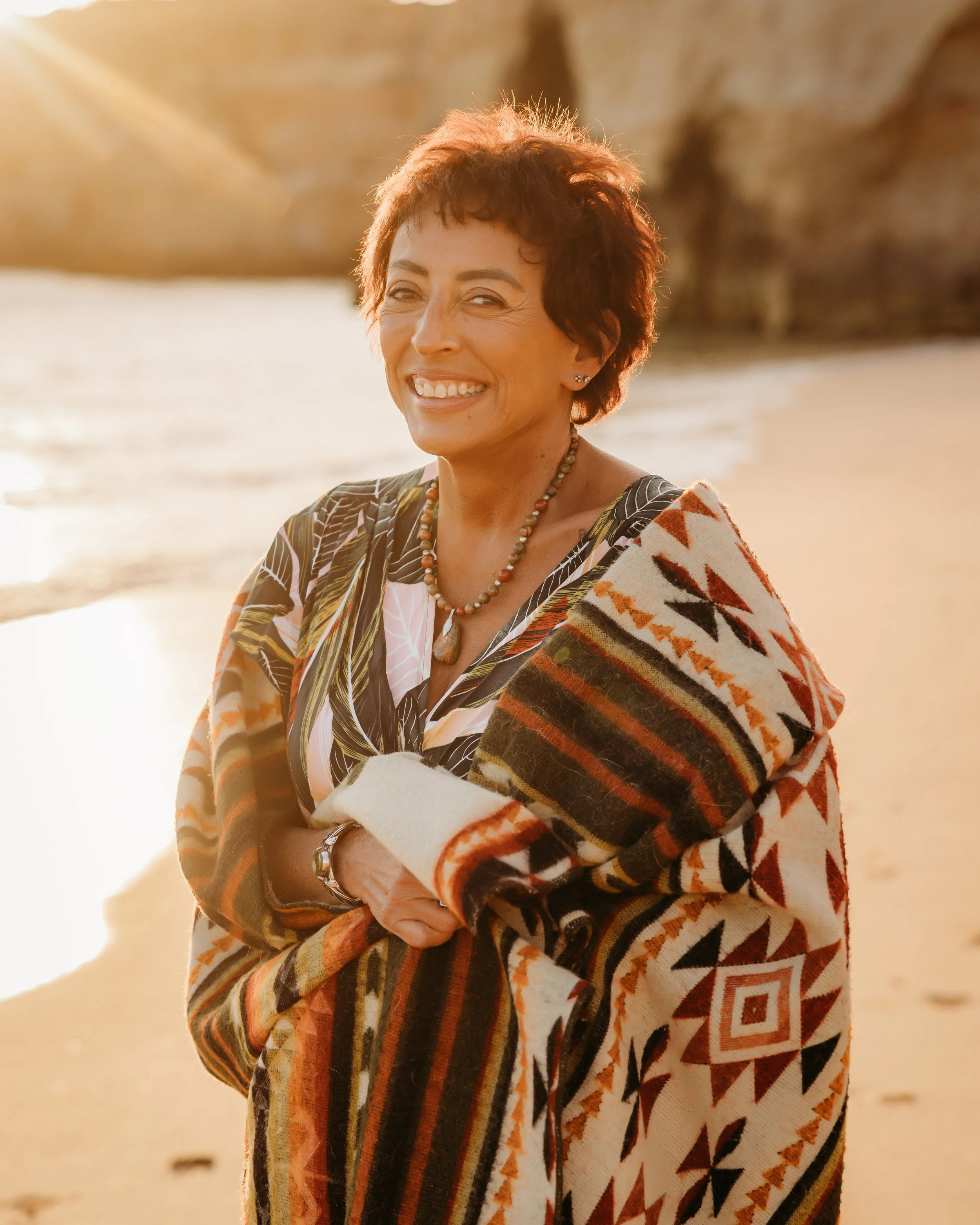 A woman with short curly hair smiling at sunset on a beach, wearing a patterned wrap with earthy tones, a beaded necklace, and bracelets.