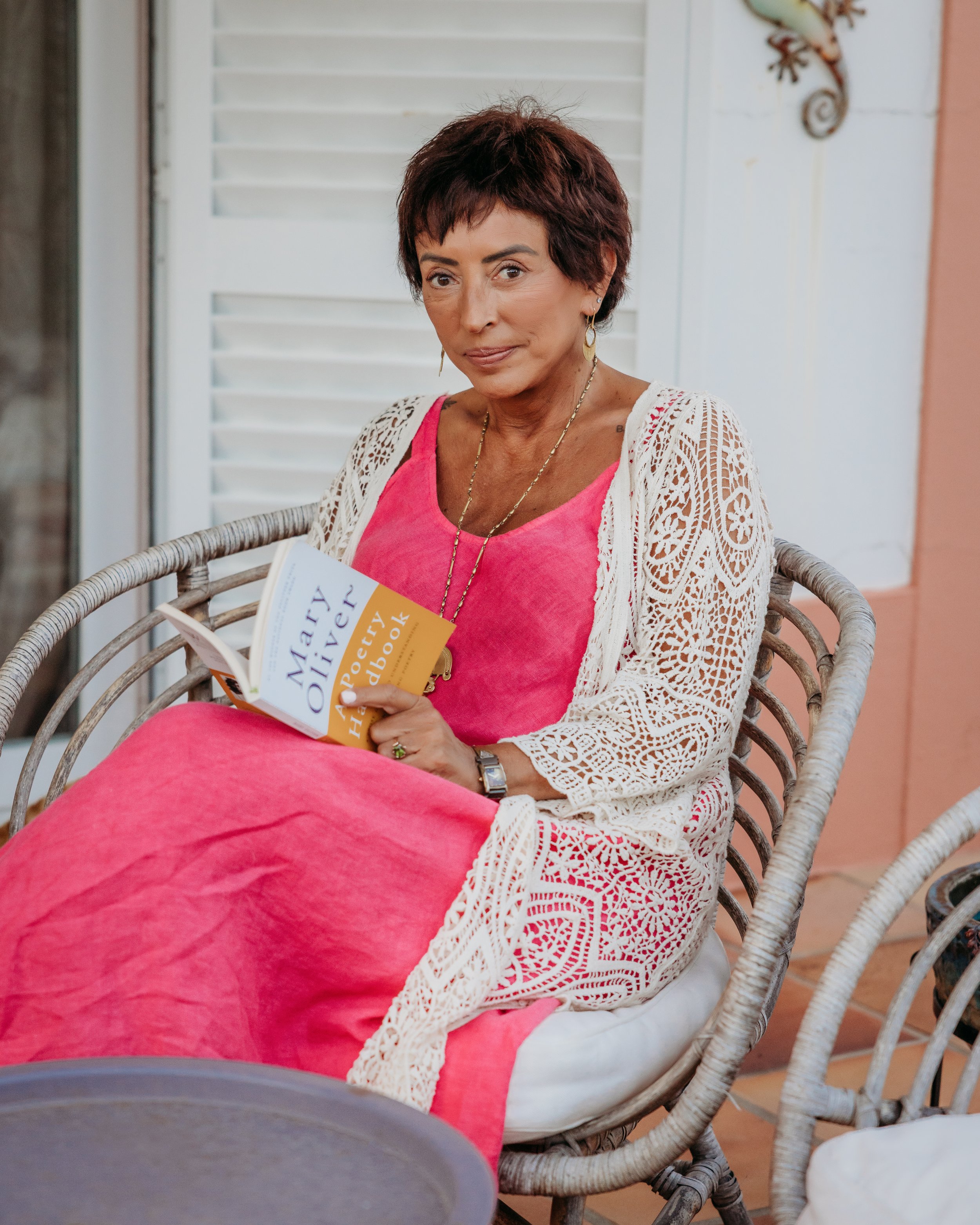 A woman with short dark hair wearing gold earrings, a gold necklace, and a white crochet cardigan over a pink top, sitting on a rattan chair, holding a paperback book titled 'Marry Me' with the subtitle 'A Novel of Love and Family.' She is seated outside near a white louvered door and a pink wall, with a decorative lizard sculpture on the wall behind her.