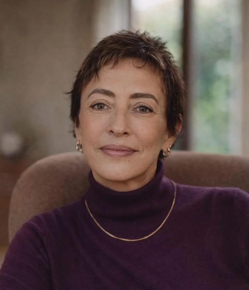Close-up of a woman with short brown hair, wearing a dark purple turtleneck, earrings, and a gold necklace, sitting indoors with a window in the background.