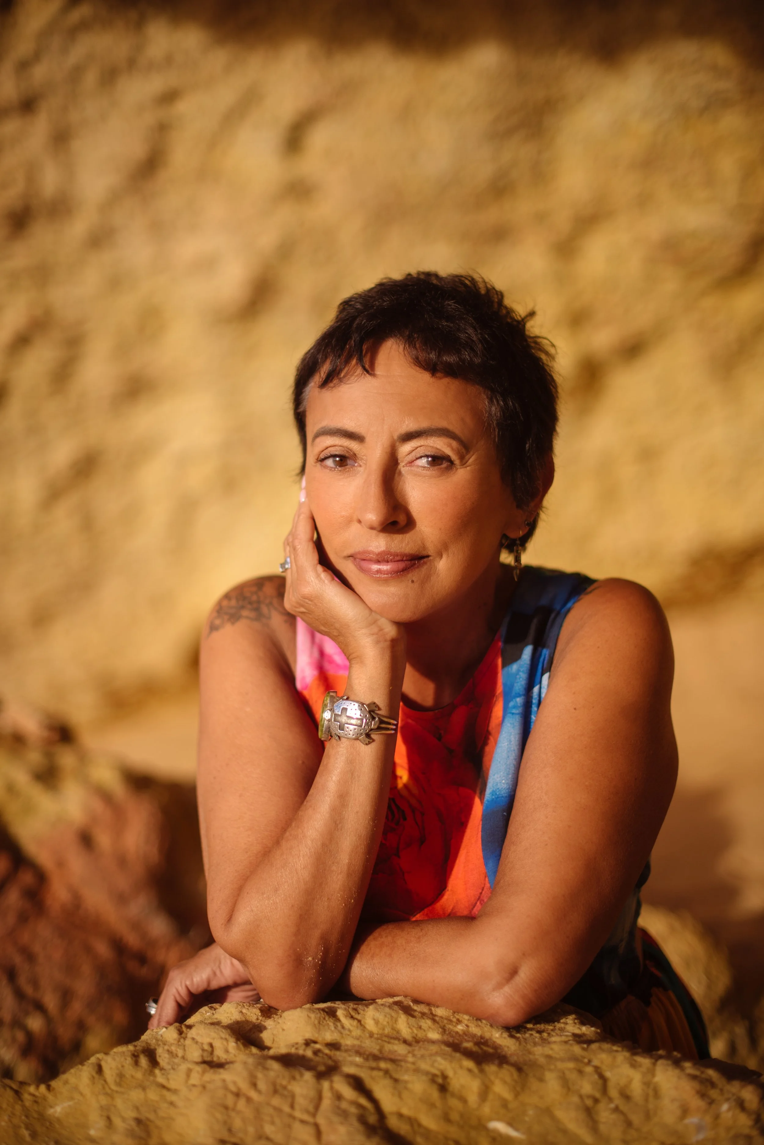 A woman with short dark hair, wearing a colorful sleeveless top, resting her head on her hand and smiling in front of a yellowish textured background.