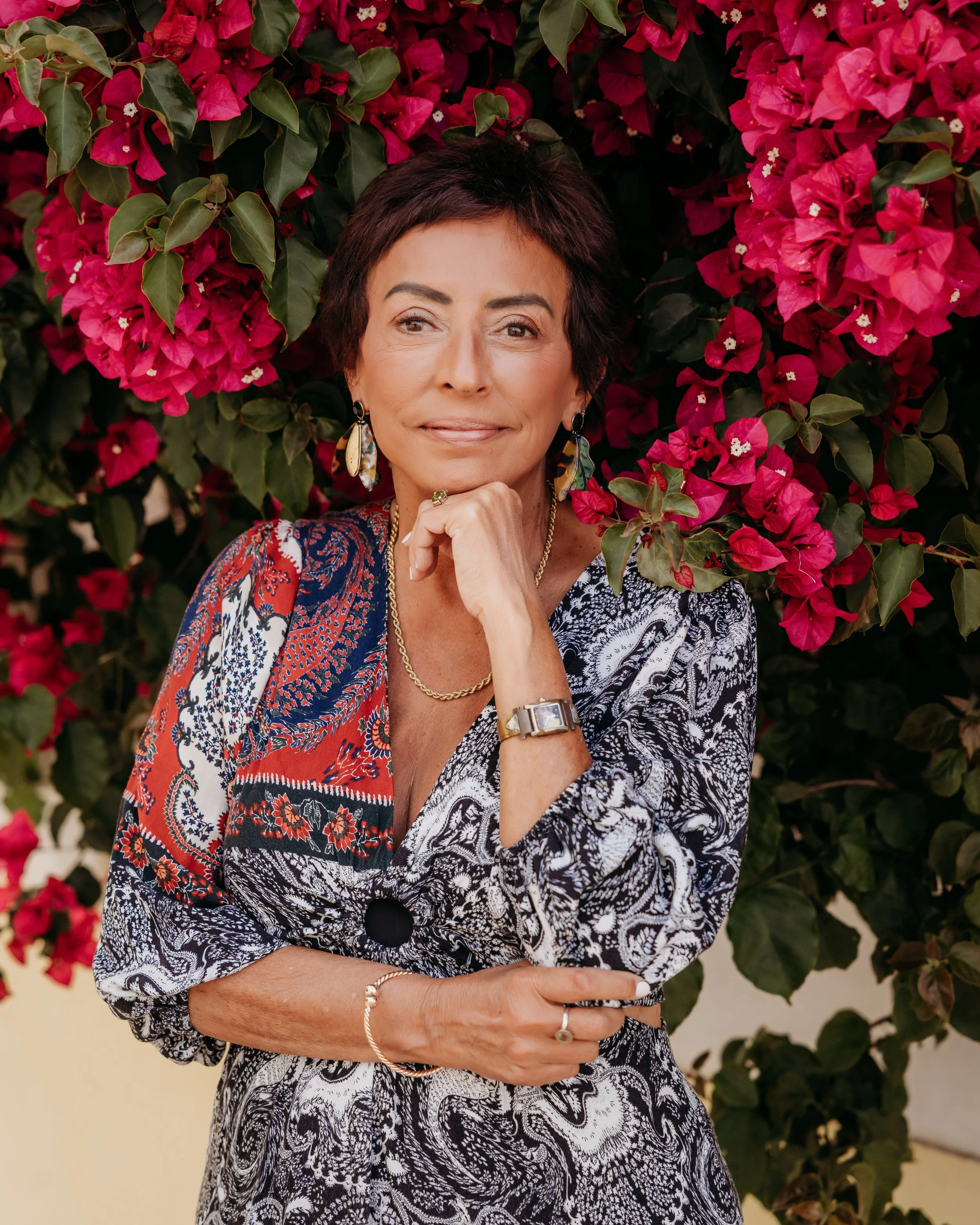 A woman with short dark hair, dressed in a colorful patterned top, wearing earrings, a watch, and rings, standing in front of a wall of pink and red bougainvillaea flowers.