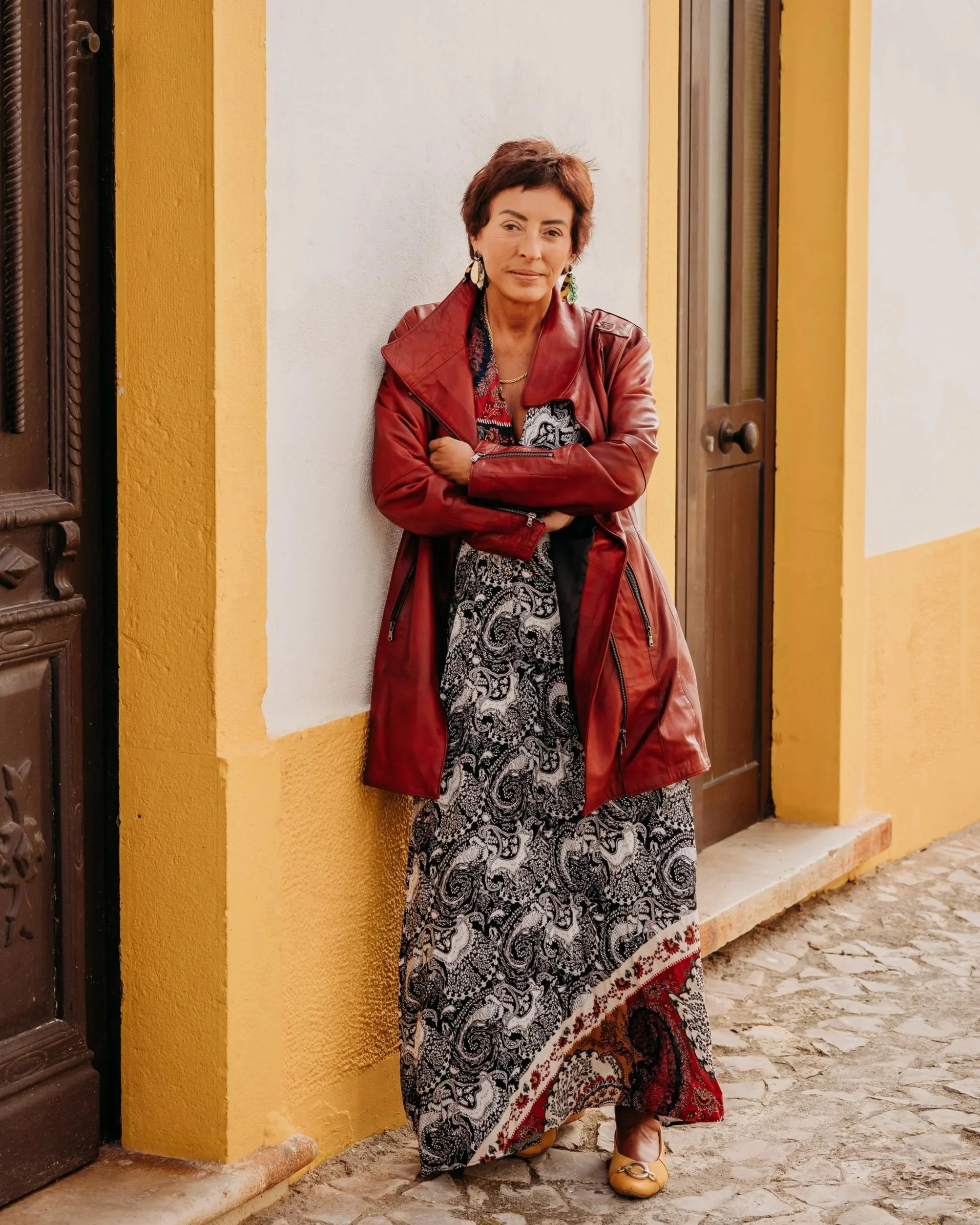 A woman standing outdoors against a yellow and white wall, wearing a red leather jacket, a patterned long skirt, beige shoes, and large earrings.