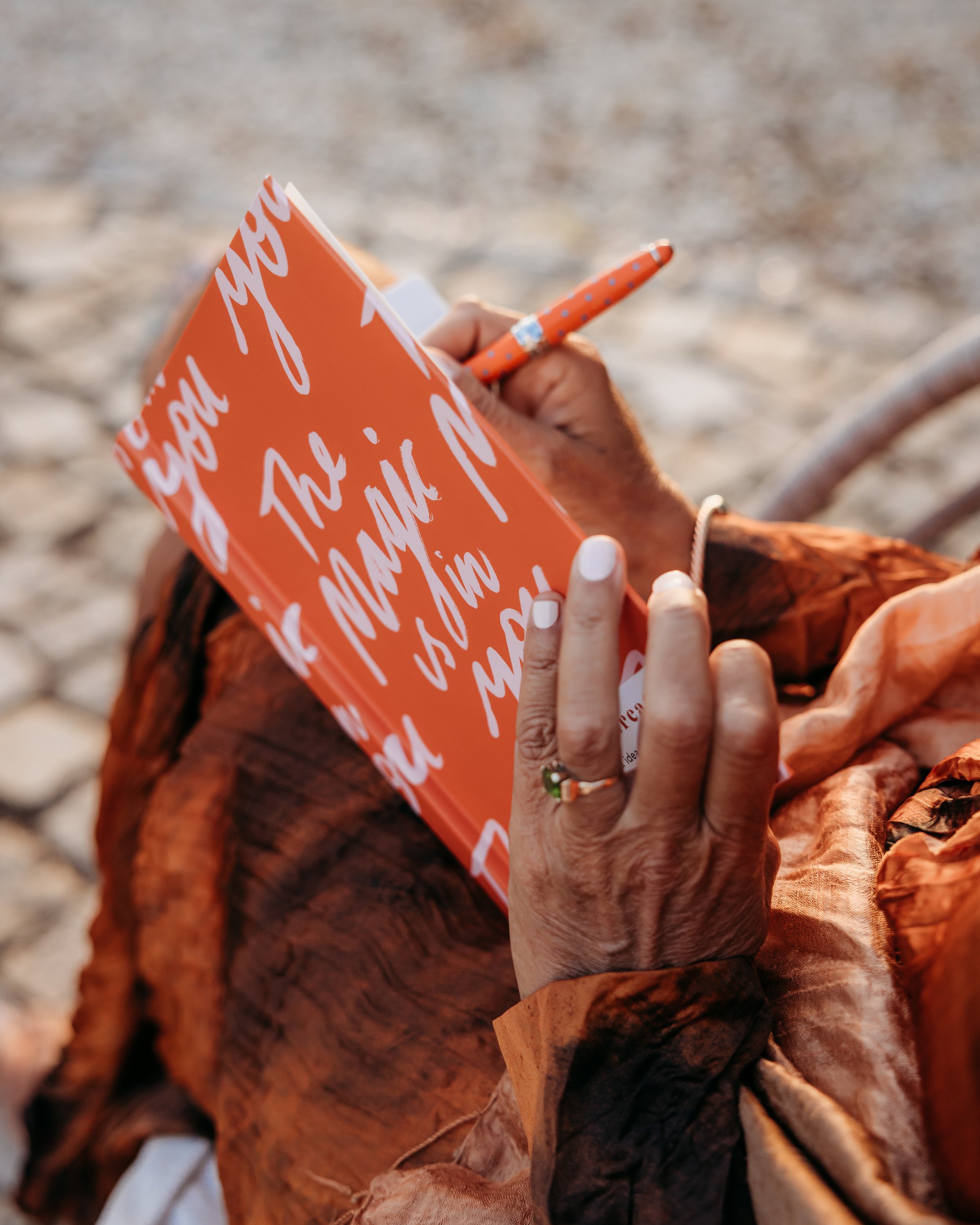 Close-up of a person's hands writing in an orange notebook with white cursive text. The person has a ring on their ring finger and is wearing a patterned, rust-colored garment. The background appears to be a gravel or sandy surface.