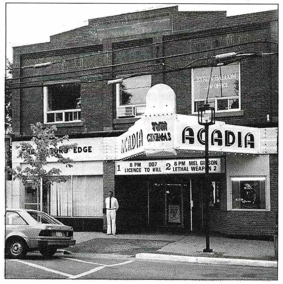 A black and white photo of an old cinema building named 'Acadia.' The marquee displays movies with showtimes and ratings, including 'Licence to Kill' and 'Lethal Weapon 2.' A man in a white suit stands outside the entrance, next to a parked car.