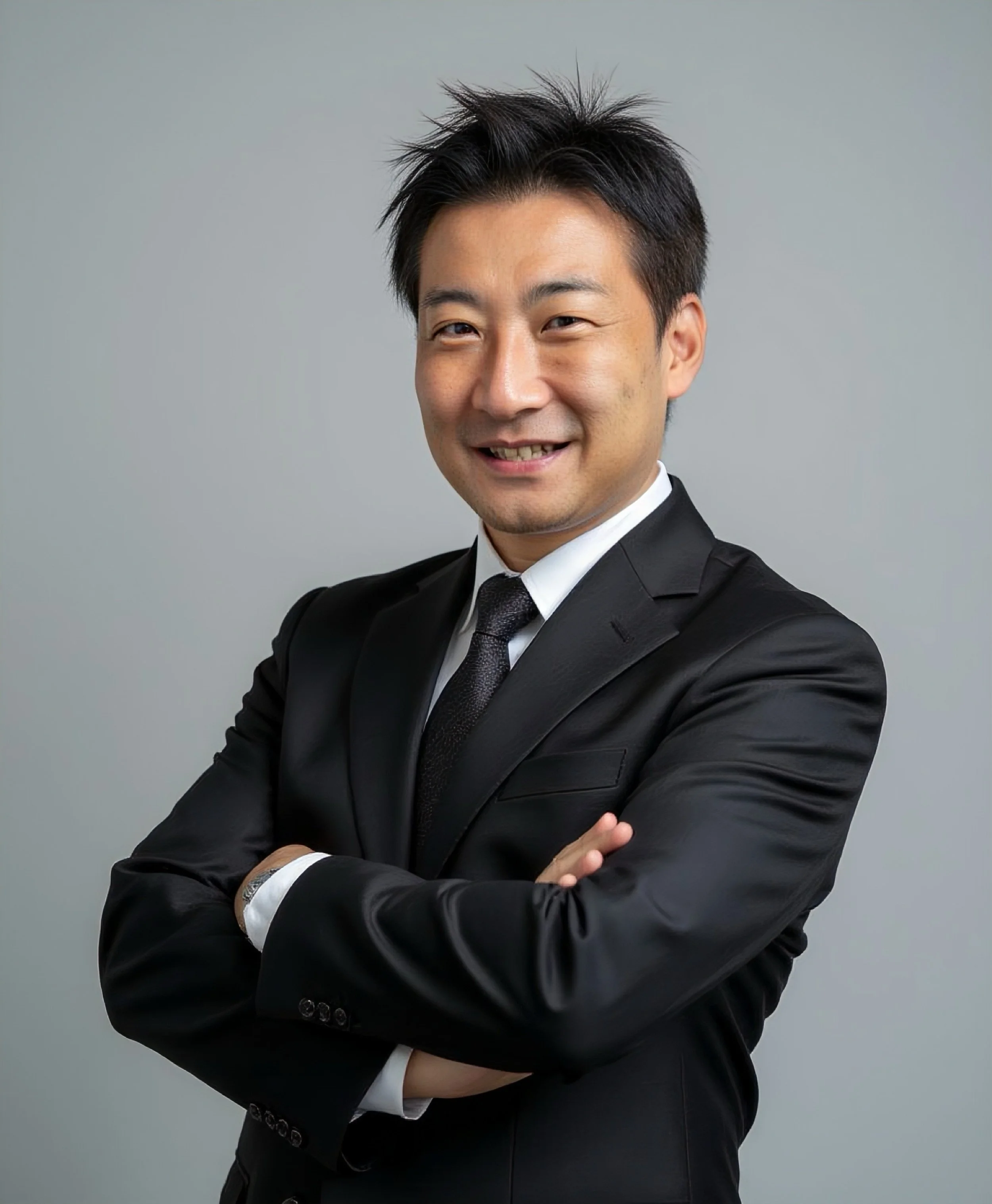 A smiling Asian man in a black suit and tie, crossing his arms in front of a plain gray background.