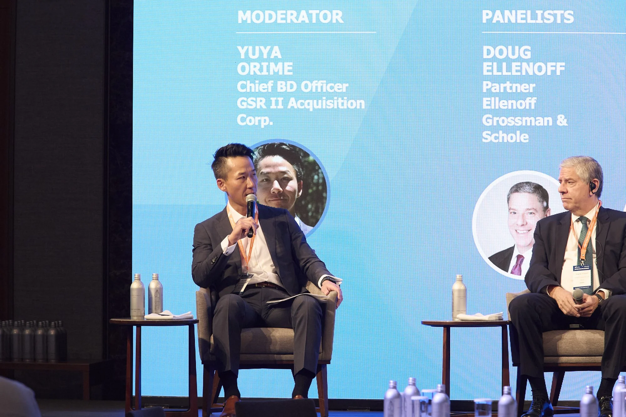 Two men in suits participating in a panel discussion on stage, seated on chairs with small tables holding water bottles beside them. Behind them, a large blue presentation screen displays text and photos, with one man speaking into a microphone and t
