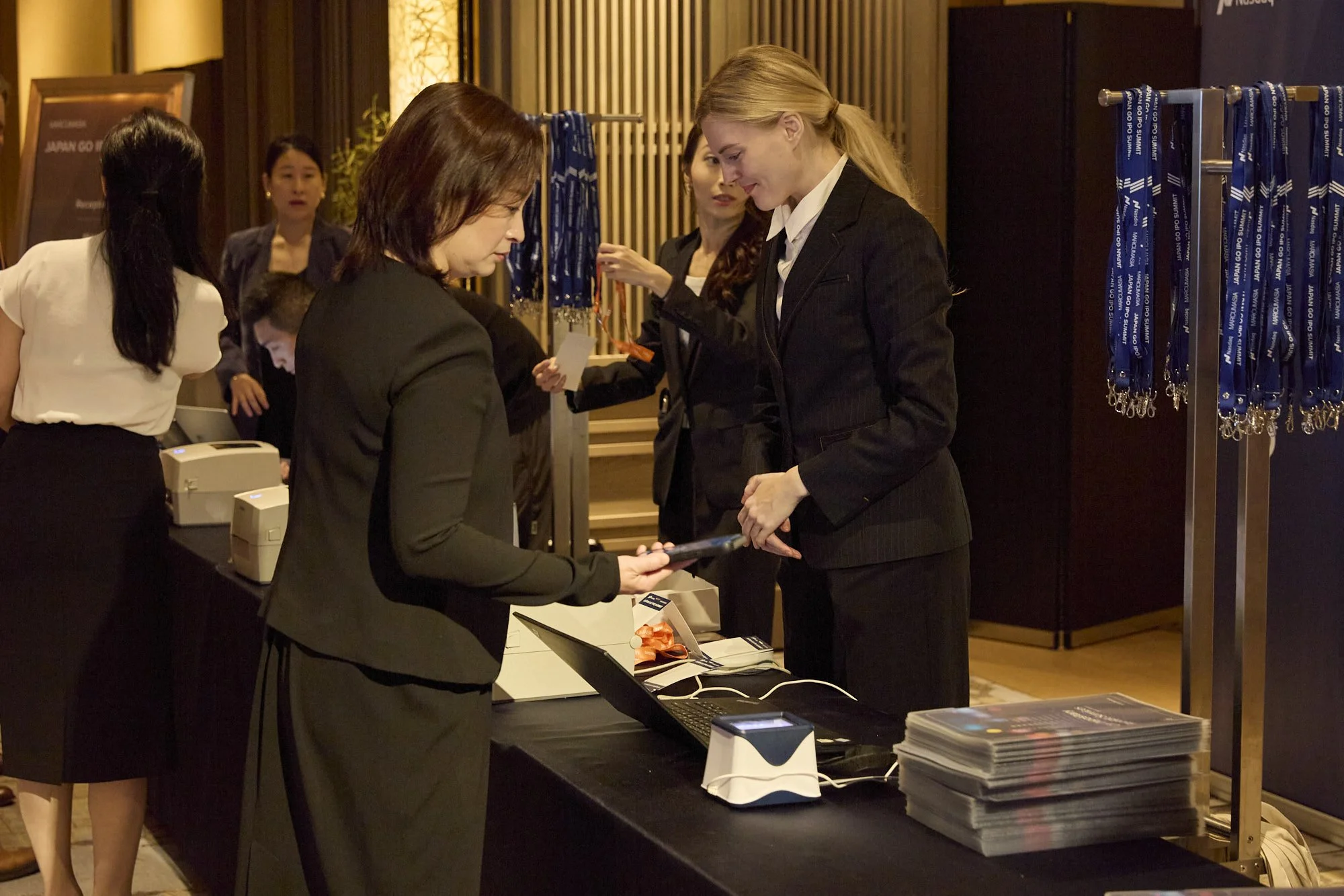 Business event with multiple women at registration desk, exchanging documents, surrounded by event materials and lanyards.