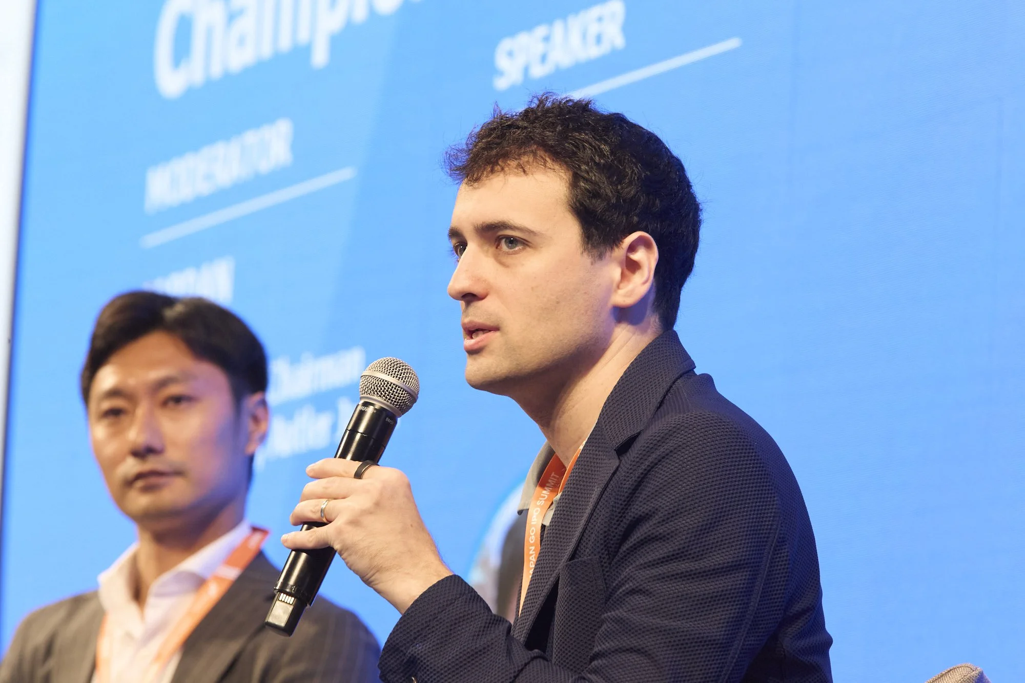 A man with dark curly hair speaking into a microphone during a panel discussion or conference, with a blue backdrop and partial view of another panelist to his left.