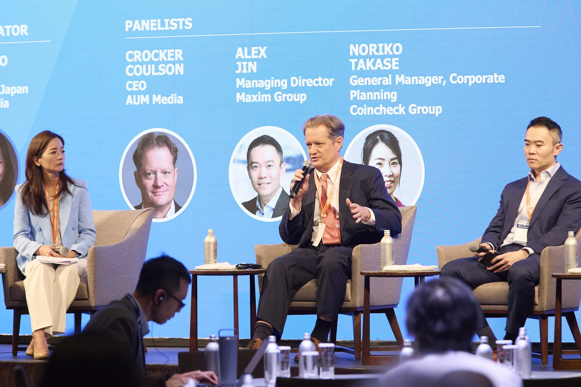 Panel discussion at a conference with four speakers seated on stage, microphone in hand, and a blue background displaying panelist names and titles.