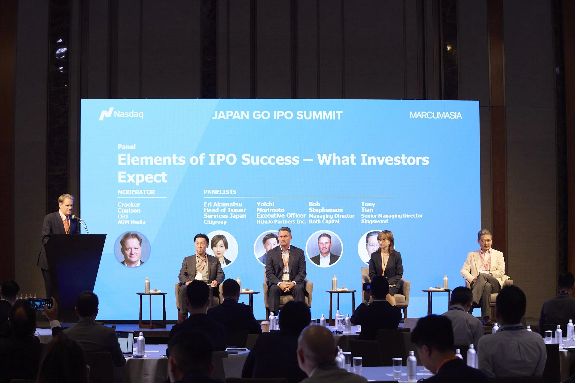 A panel discussion at the Japan GO IPO Summit, with six panelists seated on stage, a large blue screen behind them displaying the event title and speaker names, and an audience in attendance.