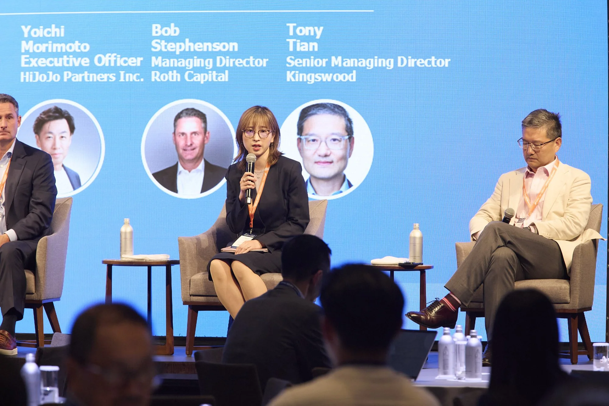 A panel discussion with five individuals on stage in front of a large blue screen displaying their names and titles. The woman in the center is speaking into a handheld microphone, wearing glasses and a black outfit. She is seated on a beige chair wi