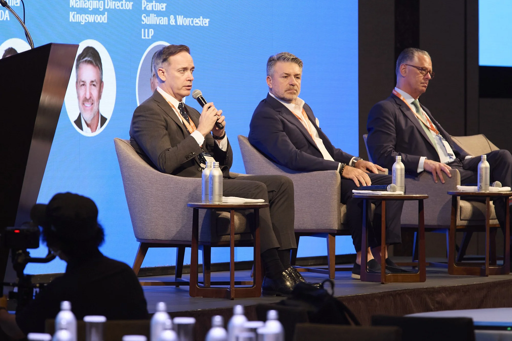 Four men sitting on chairs on a stage at a conference or seminar, with water bottles on small tables in front of them, and a large blue screen behind displaying text and photos.