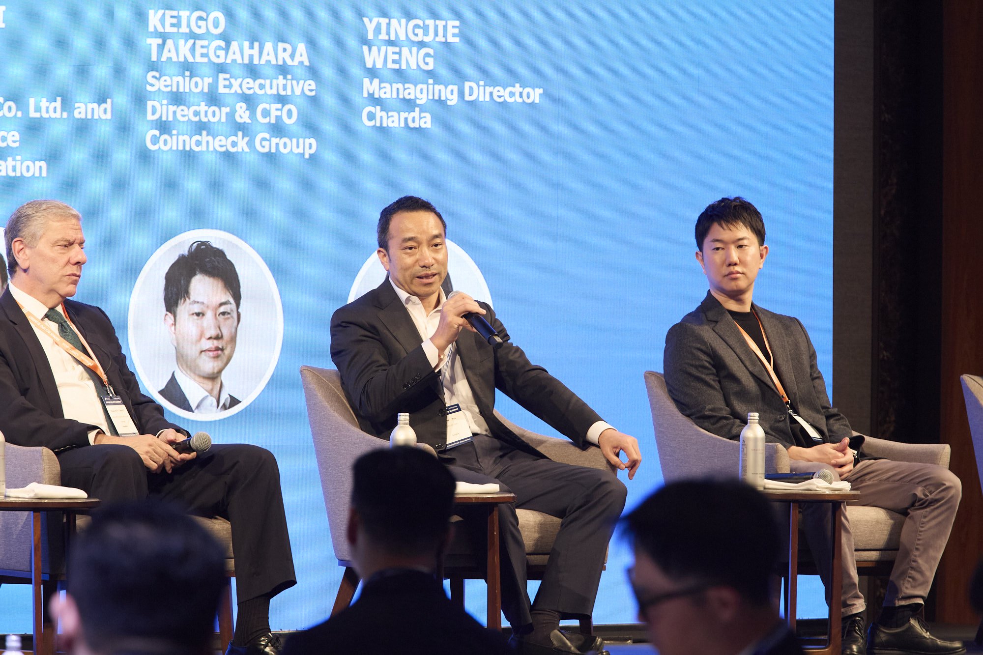 Four men participating in a panel discussion at a conference, sitting on chairs with a blue backdrop displaying their names and titles.