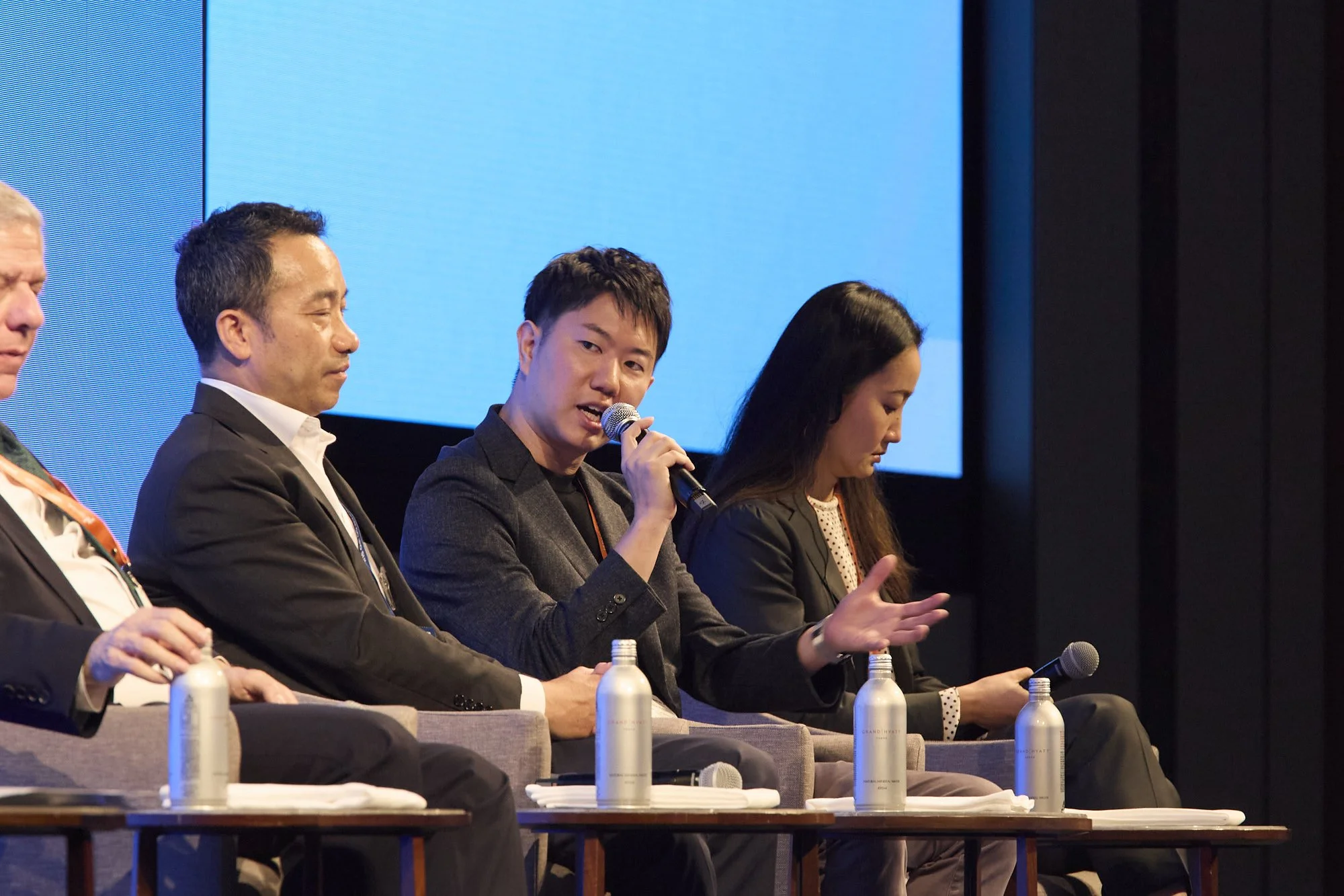 Panel of five diverse individuals seated at a table on stage, participating in a conference or panel discussion, with blue background and microphones in front of them.