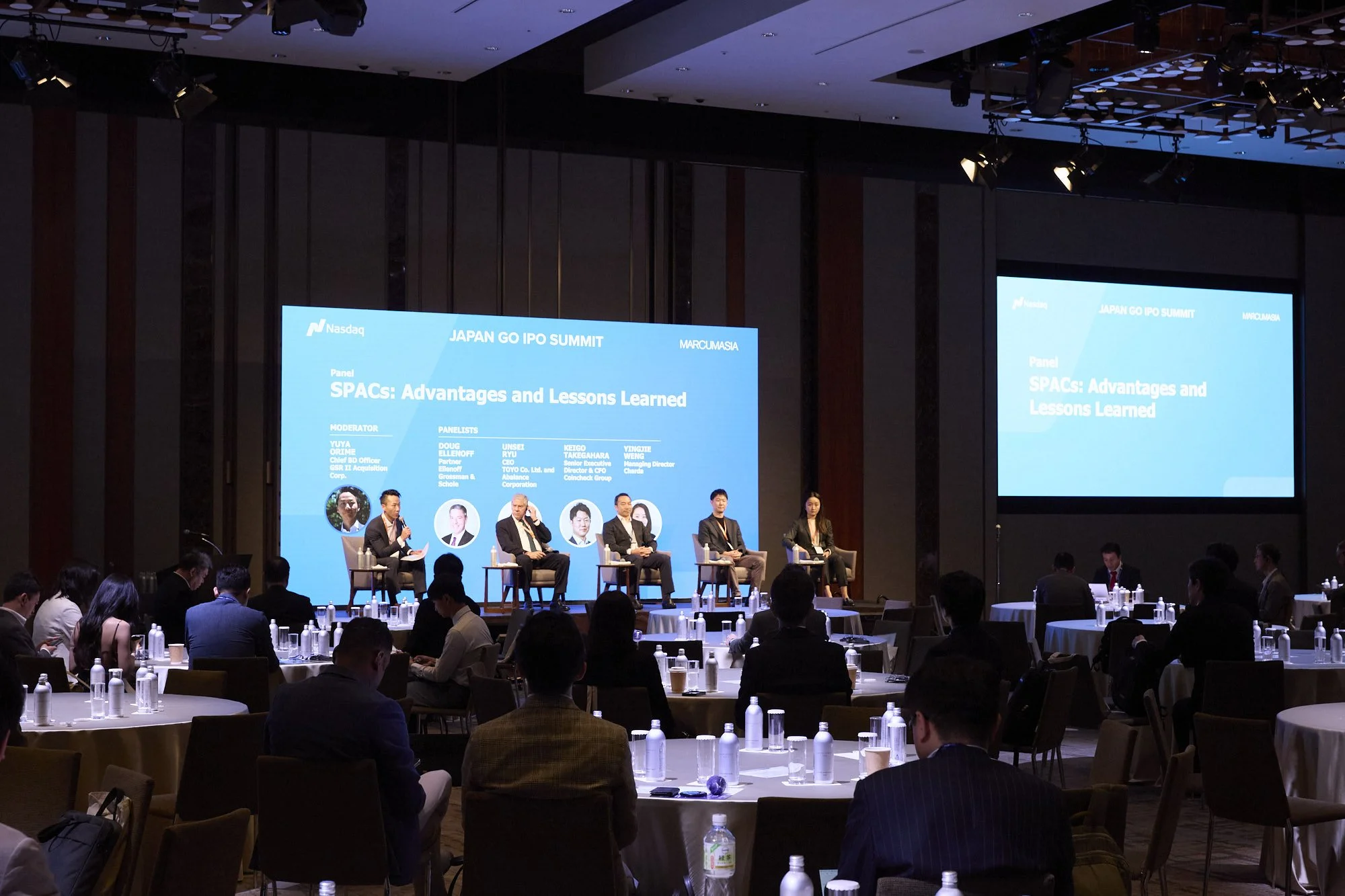 A conference room with a panel of speakers seated on stage, discussing at the Japan GO IPO Summit, with audience members seated at round tables. Large screens display the event title, panel topic, and panelists' information.