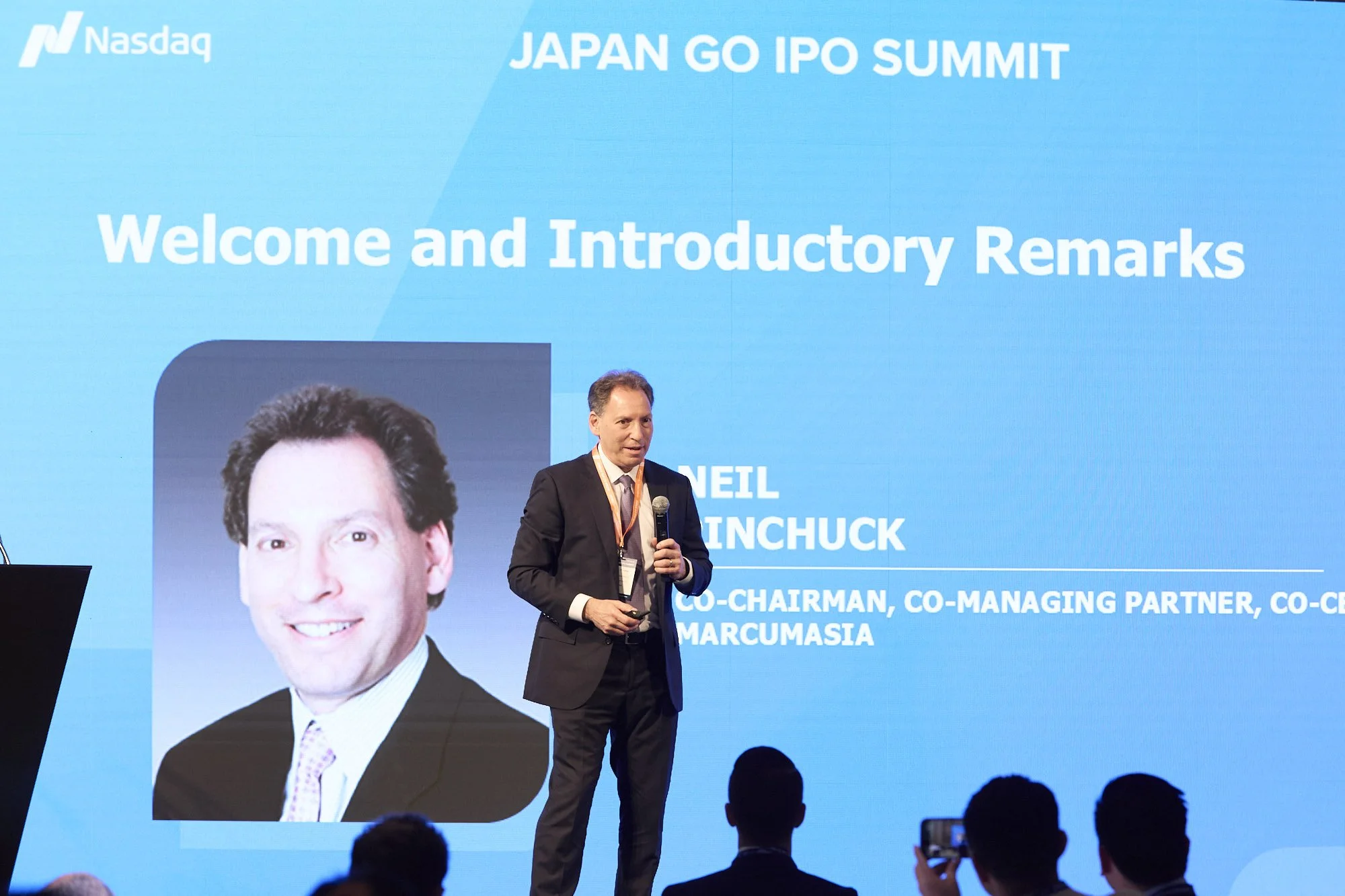 A man in a suit speaking at a conference on stage with a large blue screen behind him. The screen displays the event name, 'JAPAN GO IPO SUMMIT,' and a welcome message. The screen also features a corporate headshot of another man, along with his name