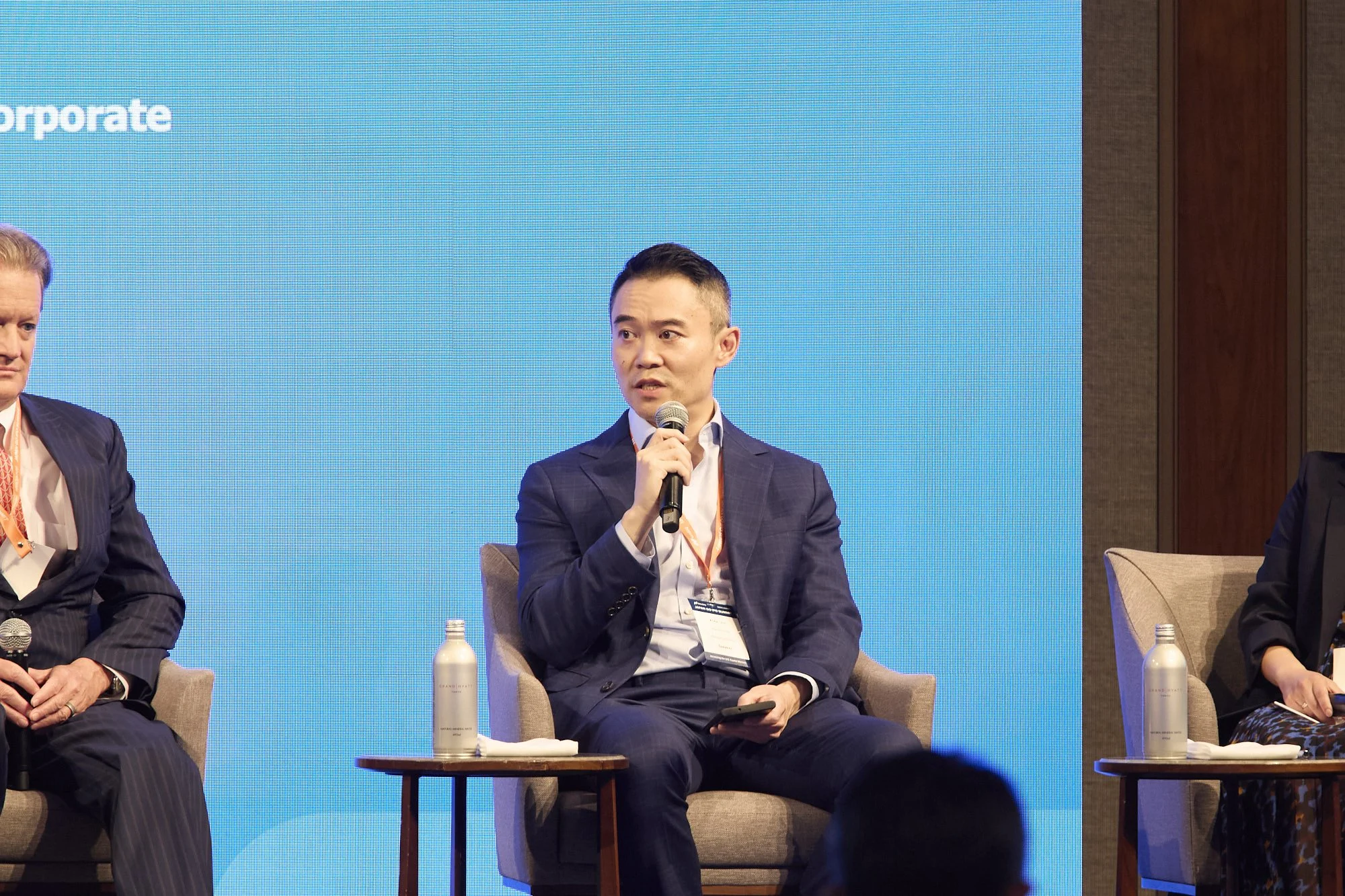 A man in a dark suit holding a microphone and speaking at a panel discussion, seated in a beige chair with a water bottle on a small table in front of him, with a blue backdrop and other panelists partially visible.