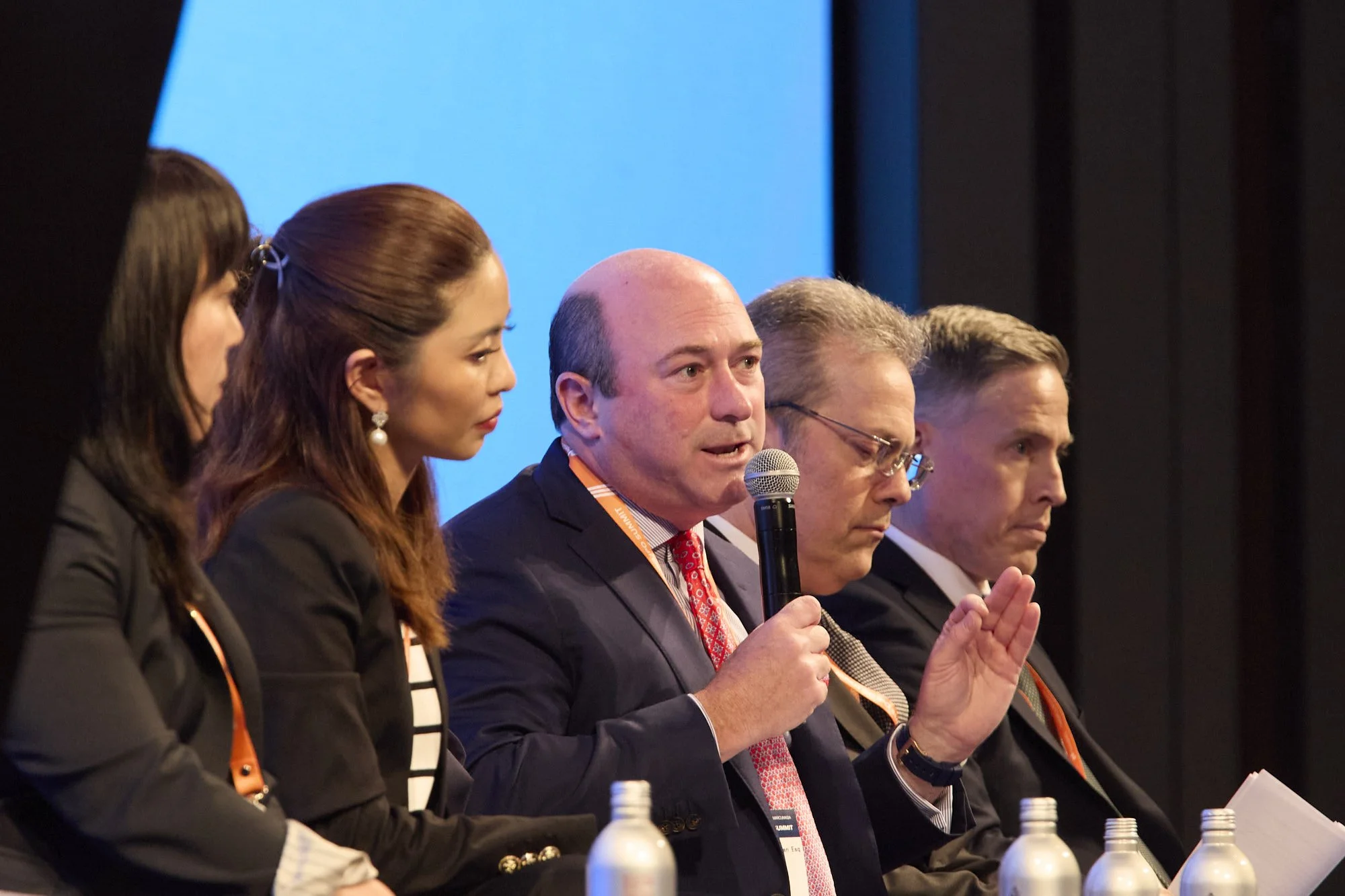 A panel of five people sitting at a table during a conference or discussion, with a blue background behind them, and water bottles in front of each person.