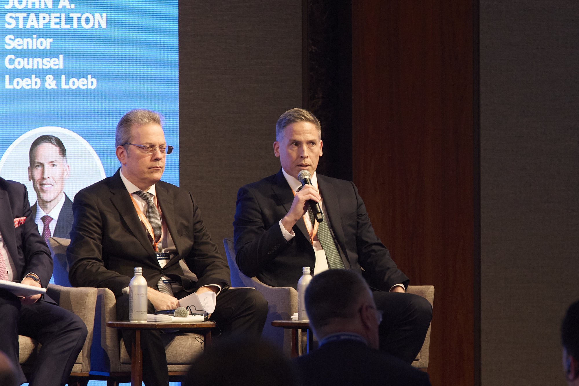 A panel of three men in suits seated at a conference, with one man speaking into a microphone. Behind them is a digital screen displaying a blue background and text including "John A. Stapleton," "Senior Counsel," and "Loeb & Loeb." There are water b