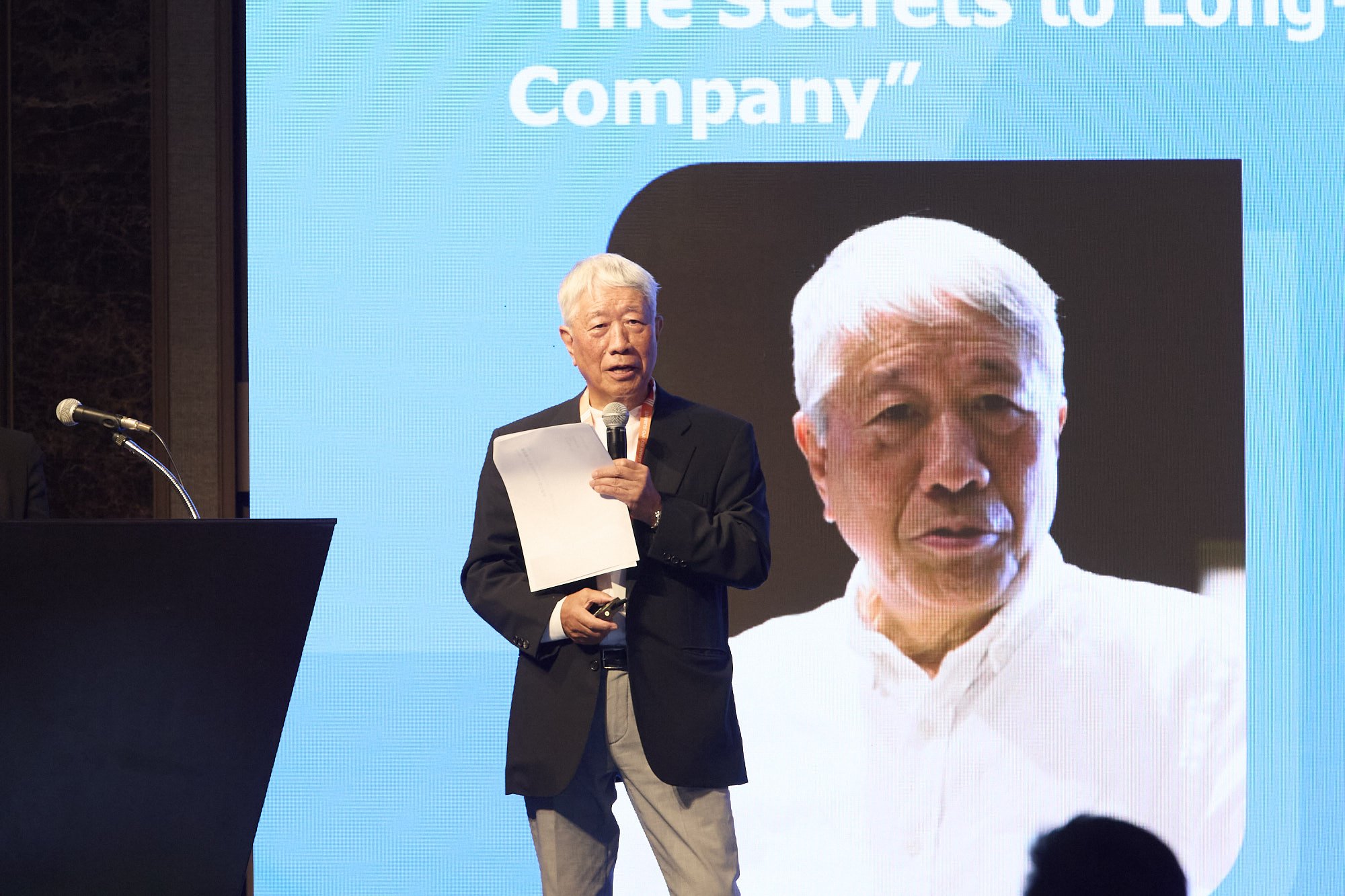 An elderly man with gray hair in a suit holding a microphone and some papers giving a presentation on stage. Behind him is a large screen displaying a photo of another elderly man with white hair and a serious expression. The background of the slide 