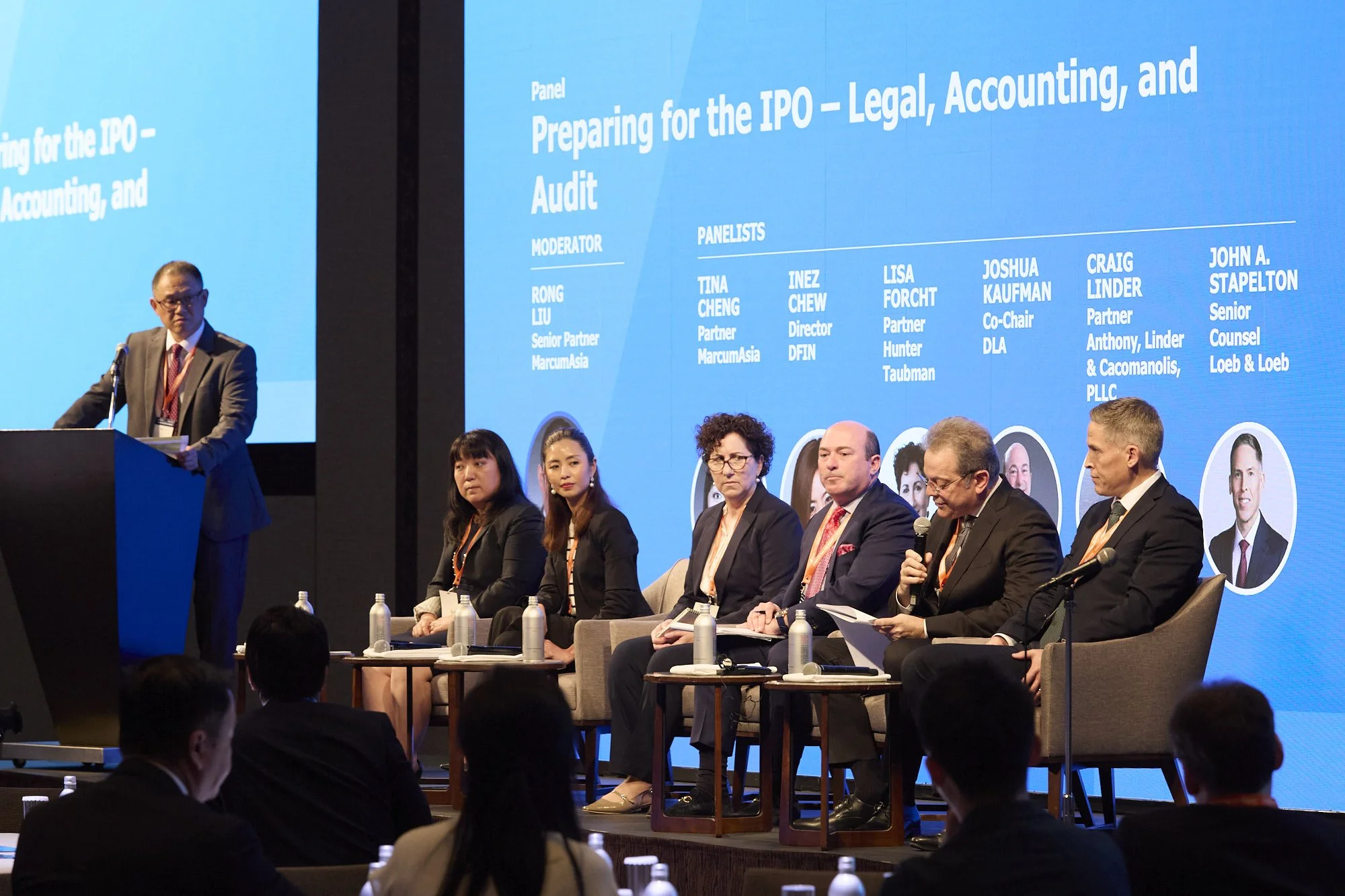 A panel discussion at a conference with seven individuals seated on stage, five women and two men, with water bottles in front of them. A man in a suit stands at a podium on the left. Behind them is a large blue screen displaying text about preparing