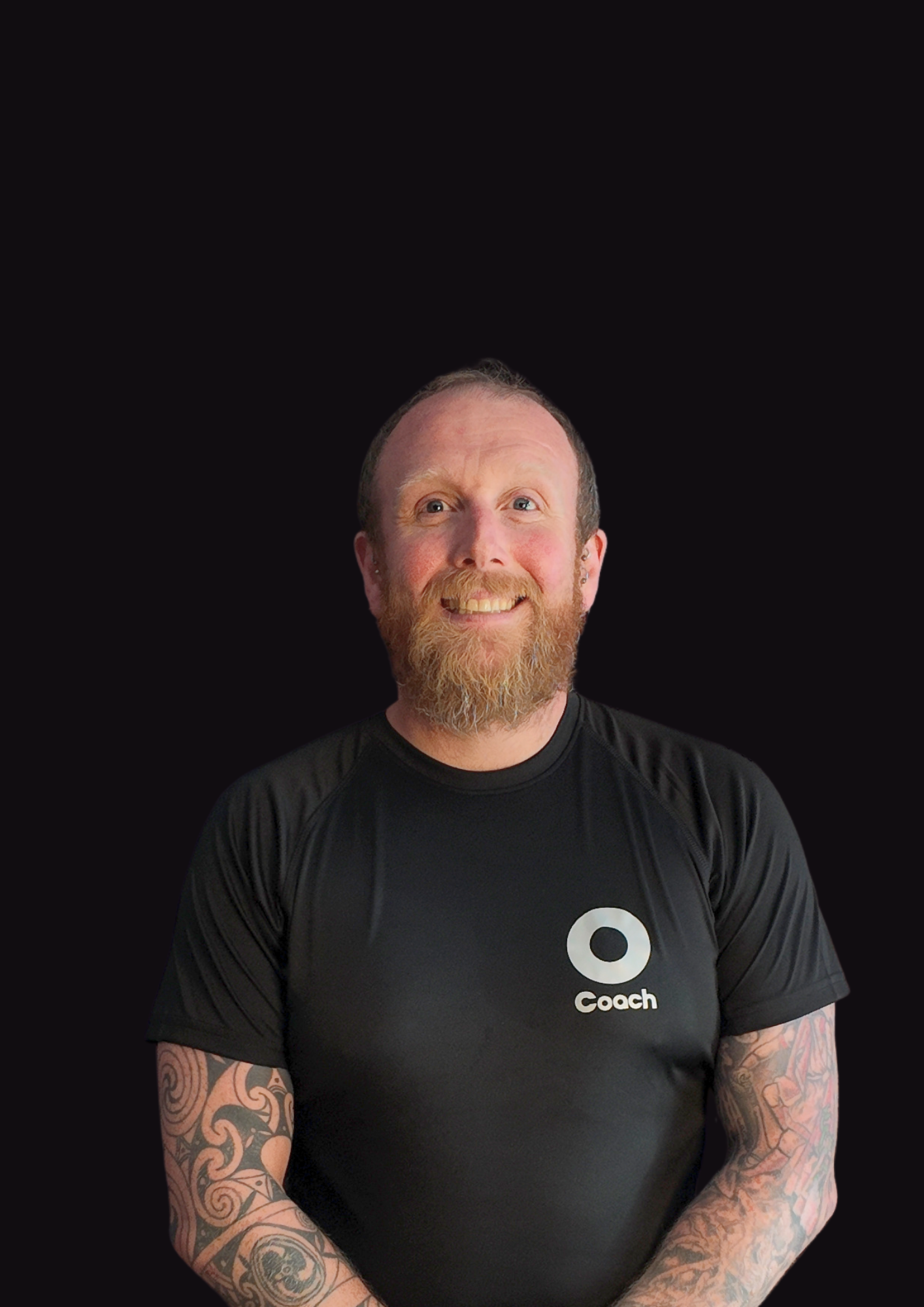 A smiling man with a beard and tattoos on his arms, wearing a black t-shirt with the word 'Coach' and a circular logo, standing against a black background.
