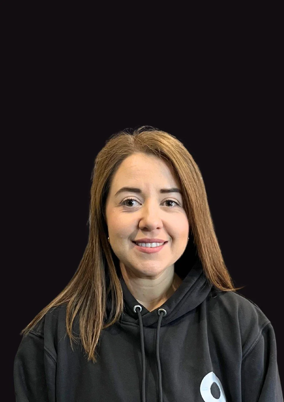 A woman with brown hair smiling, wearing a black hoodie with a white circular logo, against a plain black background.
