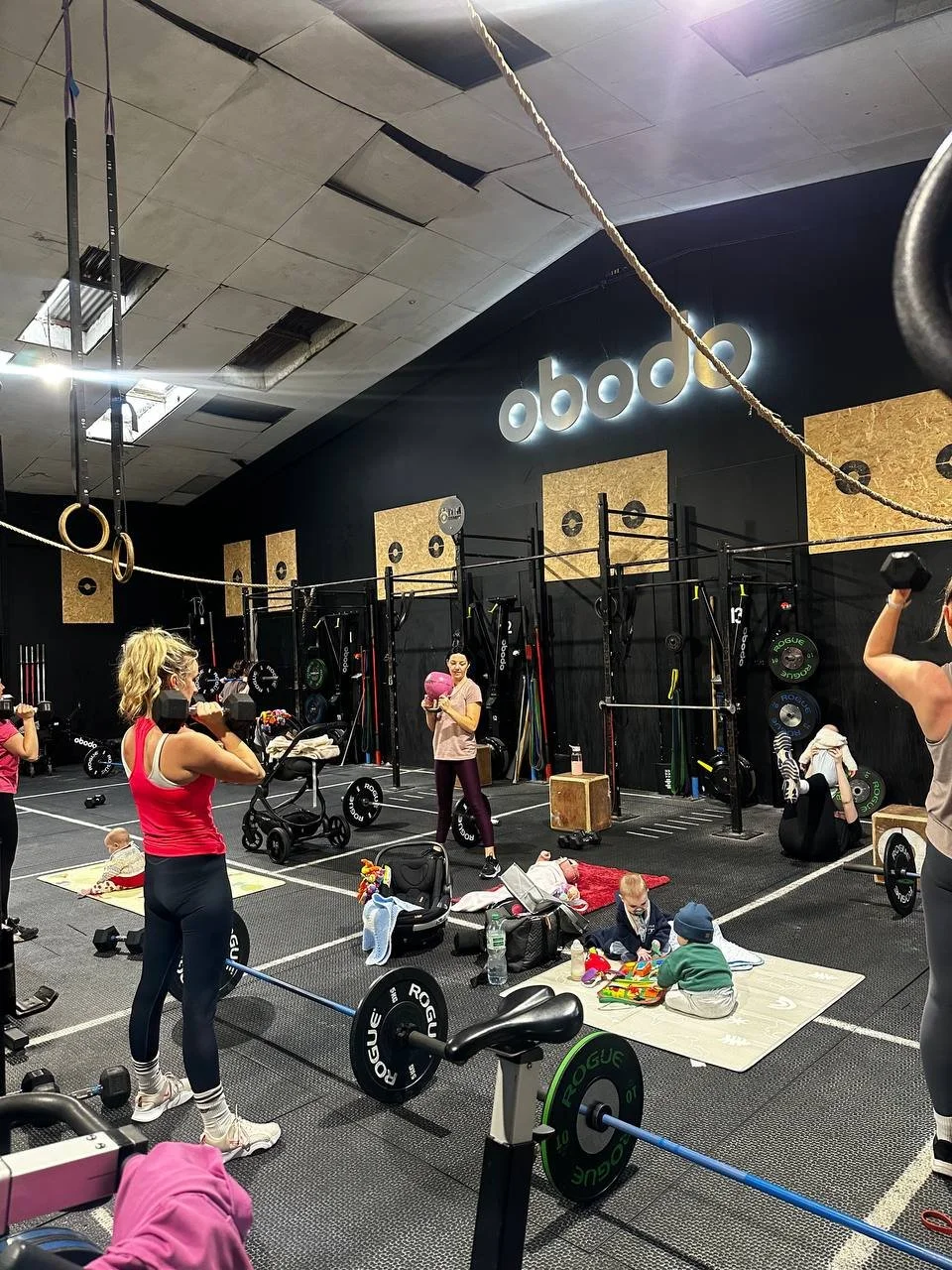 People working out with weights and doing activities in a gym with kids present, gym equipment, and the 'obod' logo on the wall.