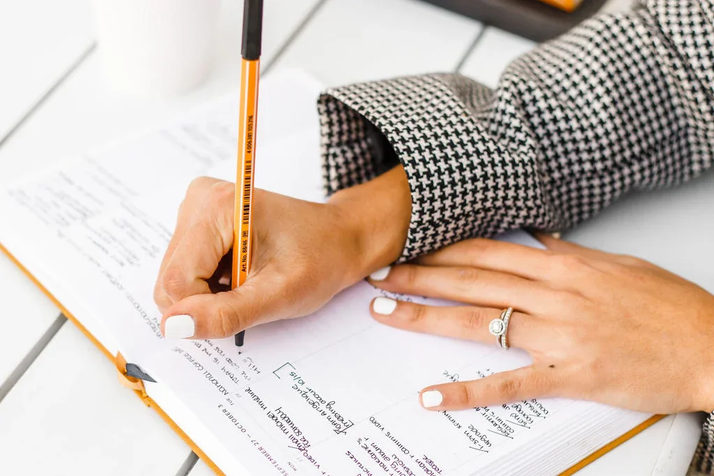 A woman writes in a notebook with a pen, resting her other hand on the table. She wears a houndstooth blazer and has a ring on her finger.