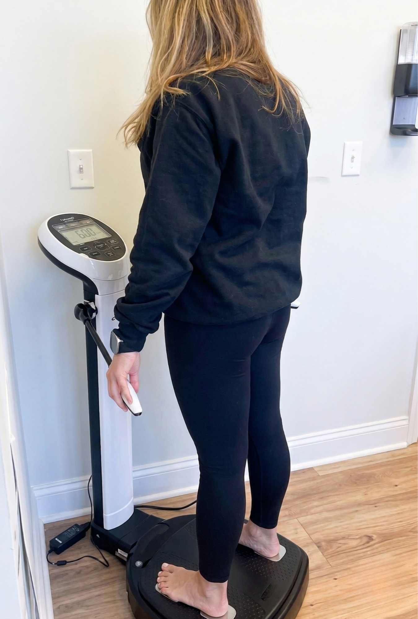 Woman standing barefoot on a digital scale, holding a remote control, in a room with white walls and wooden flooring, with a weight of 160 pounds displayed on the scale.